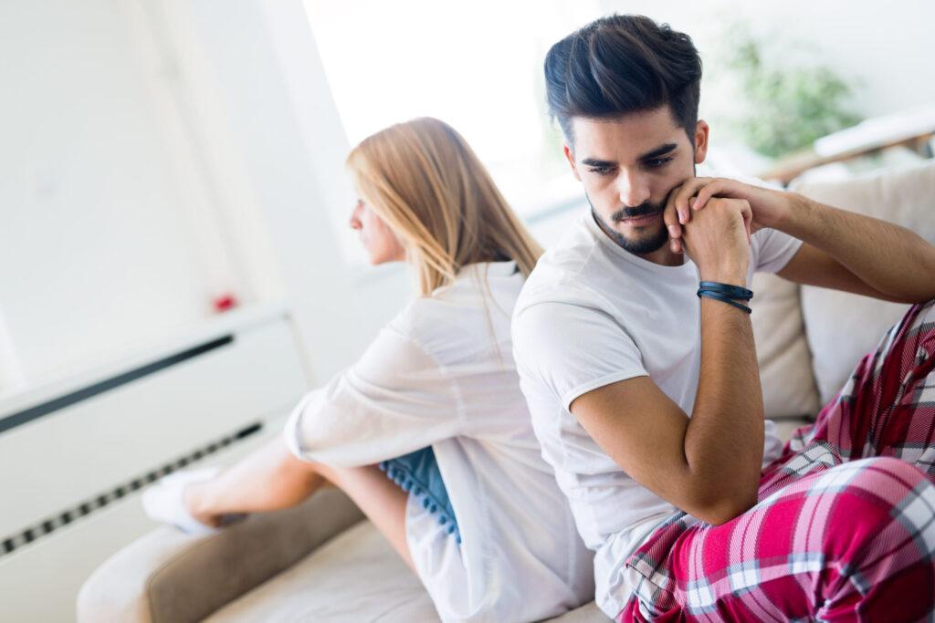 Smutna para siedząca osobno na kanapie, symbolizująca kryzys w małżeństwie spowodowany impotencją i zazdrością